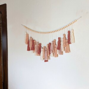 May include: A wall hanging featuring a series of tassels in shades of cream, peach, and terracotta. The tassels hang from a string of wooden beads, creating a decorative and textured piece. The wall hanging is mounted on a white wall.