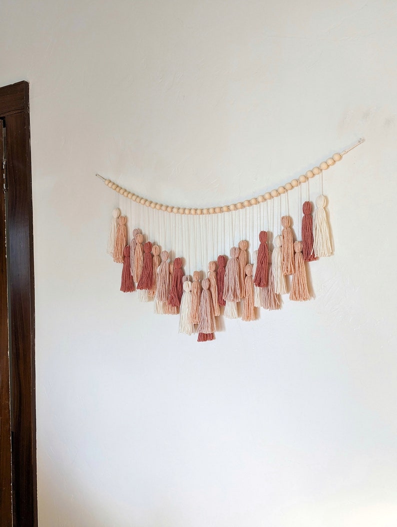 May include: A wall hanging featuring a series of tassels in shades of cream, peach, and terracotta. The tassels hang from a string of wooden beads, creating a decorative and textured piece. The wall hanging is mounted on a white wall.