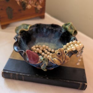 May include: A dark blue ceramic bowl with a wavy, uneven rim featuring sculpted decorative elements including a brown frog, a red bud, and green leaves. The bowl contains a strand of white pearls resting inside the basin on a dark surface.