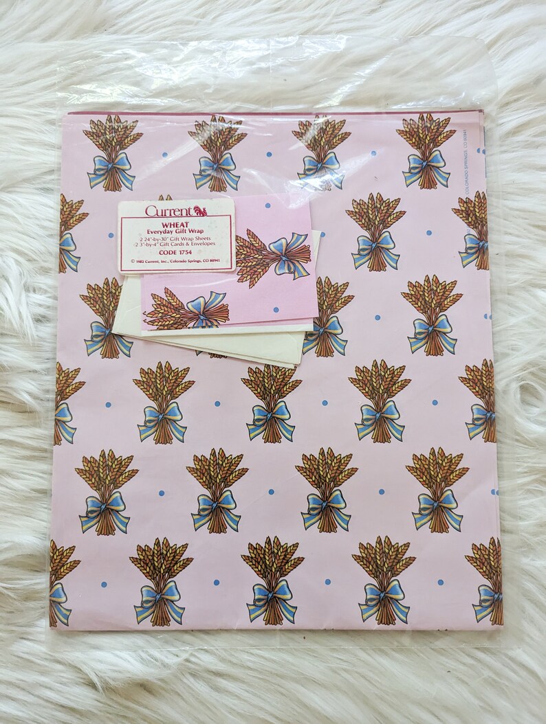 K&ouml;nnte beinhalten: Eine Packung pinkfarbenes Geschenkpapier mit Weizenmotiv, blauen Schleifen und wei&szlig;en Geschenkkarten. Die Packung enth&auml;lt 22 Geschenkpapierb&ouml;gen im Format 61 cm x 76 cm und 23 Geschenkkarten und Umschl&auml;ge im Format 7,6 cm x 10,2 cm. Der Text auf der Packung lautet "Current WHEAT Everyday Gift Wrap 22 24-by-30 Gift Wrap Sheets 23 3-by-4 Gift Cards & Envelopes CODE 1754 COLORADO SPRINGS, CO 80911"