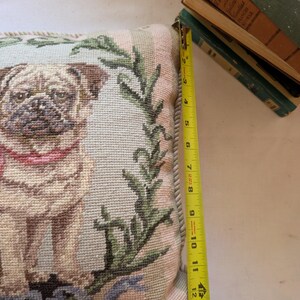May include: A square needlepoint pillow featuring a tan pug with a pink collar, surrounded by a green leafy wreath on a light blue background. A yellow measuring tape placed along the side shows the pillow height is approximately 13.5 inches.