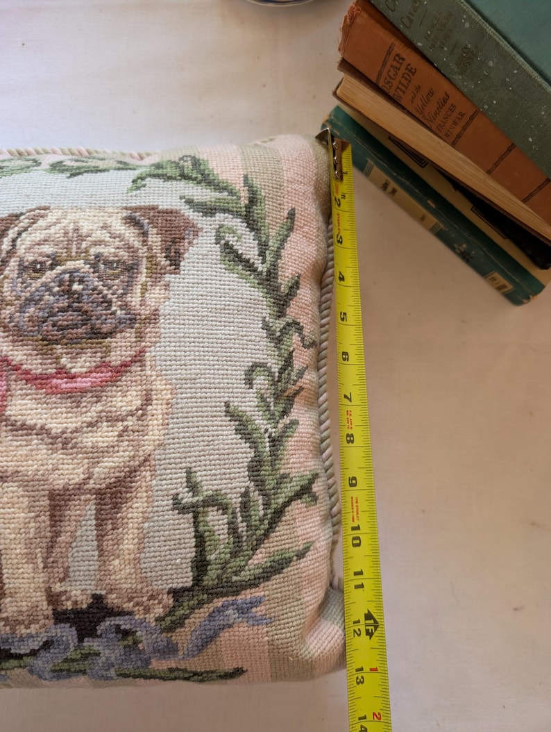May include: A square needlepoint pillow featuring a tan pug with a pink collar, surrounded by a green leafy wreath on a light blue background. A yellow measuring tape placed along the side shows the pillow height is approximately 13.5 inches.