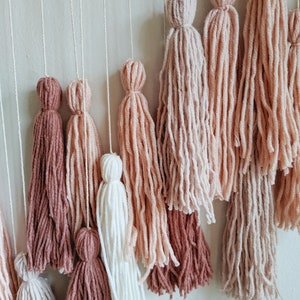 May include: A wall hanging featuring multiple yarn tassels in varying shades of pink, white, and brown. The tassels are suspended from thin white cords, creating a layered, textured design. The color palette is soft and neutral.