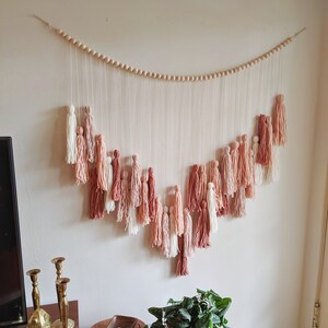May include: A wall hanging featuring a series of tassels in shades of white, pink, and terracotta. The tassels are suspended from a string of wooden beads, creating a decorative, bohemian-style piece. The wall hanging is mounted on a white wall.