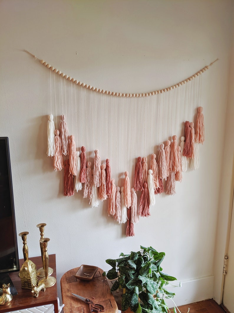 May include: A wall hanging featuring a series of tassels in shades of white, pink, and terracotta. The tassels are suspended from a string of wooden beads, creating a decorative, bohemian-style piece. The wall hanging is mounted on a white wall.