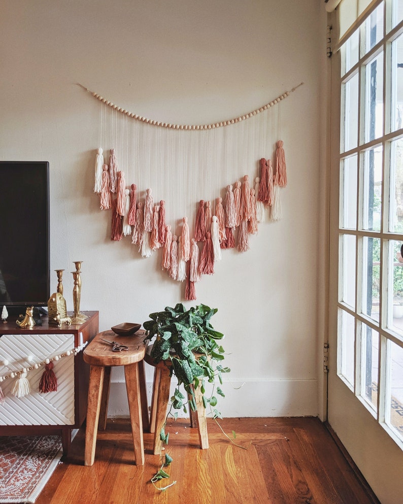 May include: A wall hanging with a wooden bead garland and cascading tassels in shades of pink, peach, and white. The tassels are suspended from strings, creating a decorative, bohemian-style accent. The wall hanging is displayed above a wooden stool and a cabinet.