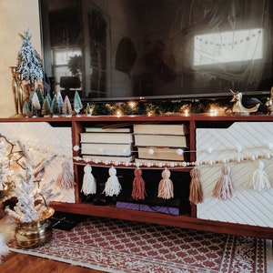 Peut inclure: Un meuble de divertissement en bois avec un motif en chevron blanc et brun sur les portes. Le meuble a un dessus plat avec une t&eacute;l&eacute;vision, une guirlande de lumi&egrave;res et divers objets d&eacute;coratifs. Le meuble a une &eacute;tag&egrave;re avec des livres et une cha&icirc;ne de pompons blancs avec des perles en bois.