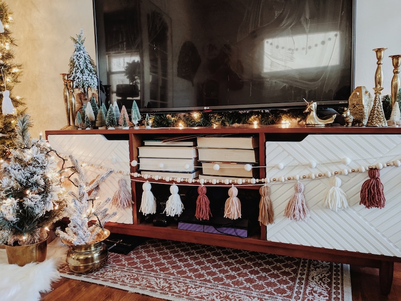 Peut inclure: Un meuble de divertissement en bois avec un motif en chevron blanc et brun sur les portes. Le meuble a un dessus plat avec une t&eacute;l&eacute;vision, une guirlande de lumi&egrave;res et divers objets d&eacute;coratifs. Le meuble a une &eacute;tag&egrave;re avec des livres et une cha&icirc;ne de pompons blancs avec des perles en bois.