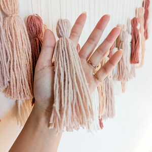 May include: A wall hanging featuring multiple yarn tassels in shades of beige, pink, and brown. The tassels are suspended from a wooden bead garland. The image shows a hand holding one of the tassels.