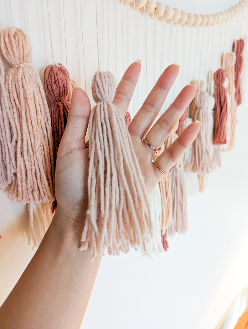 May include: A wall hanging featuring multiple yarn tassels in shades of beige, pink, and brown. The tassels are suspended from a wooden bead garland. The image shows a hand holding one of the tassels.