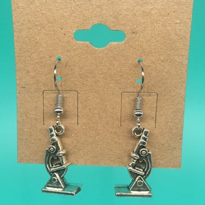 May include: A pair of silver microscope earrings. The earrings are hanging from silver hooks. The microscopes are detailed and have a silver finish.
