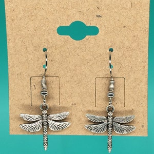 May include: Silver-toned dragonfly earrings. Each earring features a detailed dragonfly pendant suspended from a silver hook. The dragonfly has intricate wing and body detailing. The earrings are displayed on a brown cardboard earring card.