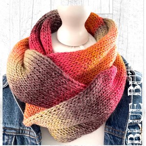 May include: A knitted infinity scarf in a gradient of colours, featuring orange, pink, and brown hues. The scarf is made from thick yarn, giving it a textured look. It is draped around a mannequin head and a denim jacket.