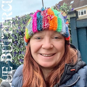 May include: A woman wearing a colourful striped crocheted hat with a light blue jacket. The hat has a variety of colours including pink, orange, green, blue, and white.