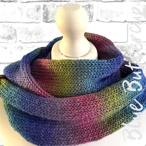 May include: A multi-coloured knitted cowl scarf, featuring shades of blue, purple, pink, and green. The scarf is draped over a wooden mannequin head, set against a white brick wall. The Blue Buttercup logo is visible.