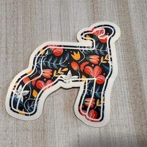 May include: A white sticker with a black outline of a sheep. The sheep is filled with a floral pattern of red, yellow, and blue flowers on a black background.
