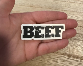 May include: A white sticker with the word "BEEF" in bold, black capital letters. The letters incorporate a cow silhouette. The sticker is rectangular and is held in a hand.