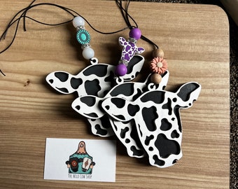 Puede incluir: Tres adornos de cabeza de vaca de madera con manchas blancas y negras, colgados de cordones negros con cuentas de colores. Los adornos están sobre una superficie de madera. El texto "The Wild Cow Shop" está en una tarjeta blanca.