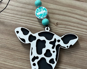 Puede incluir: Un recorte de cabeza de vaca blanco y negro con una cuerda y cuentas turquesa. La cuenta con la palabra "Mama" es turquesa con letras blancas.