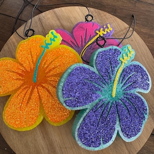 May include: Three glittery hibiscus flower ornaments in orange, pink, and purple. Each flower has a yellow stamen and a black string for hanging. The ornaments are on a wooden surface.