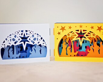 May include: Two papercraft nativity scenes, one in white with blue accents and the other in yellow with orange accents. Both scenes feature a star, palm trees, and figures of Mary, Joseph, and the baby Jesus.