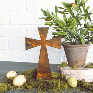 May include: A rustic metal cross with a weathered finish stands beside a potted olive tree. The tree is in a terracotta pot, set on a birch wood slice, surrounded by green moss and speckled Easter eggs. The scene evokes a sense of natural beauty and spirituality.