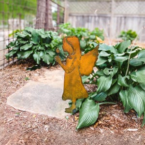 May include: A rusty metal garden sculpture of an angel holding a flower. The angel is standing on a green lawn with brown mulch.