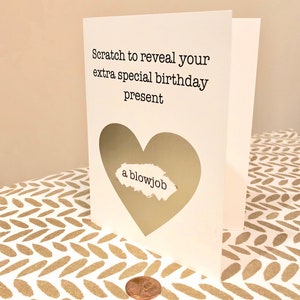 May include: A white birthday card with a heart-shaped scratch-off area. The card reads "Scratch to reveal your extra special birthday present." The revealed message says "a blowjob." A penny is next to the card.