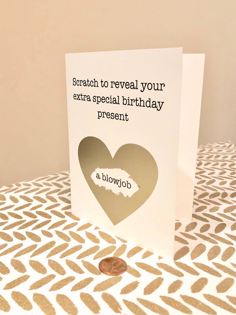 May include: A white birthday card with a heart-shaped scratch-off area. The card reads "Scratch to reveal your extra special birthday present." The revealed message says "a blowjob." A penny is next to the card.