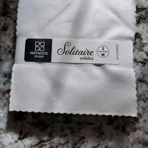 May include: A white fabric swatch with a label that reads "Maywood Studio Solitaire whites S Soft White W 42 Charms".