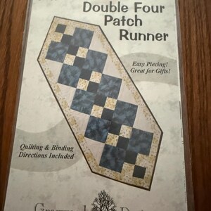 May include: A sewing pattern for a Double Four Patch Runner by Graywood Designs. The cover features a blue and cream geometric quilt runner illustration, the text GW04, Easy Piecing! Great for Gifts!, and Quilting & Binding Directions Included.