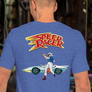 May include: Blue short-sleeve t-shirt featuring the Speed Racer logo and a graphic of Speed Racer with his car. The graphic is in yellow, red, white, and blue. The shirt is made of a soft material.