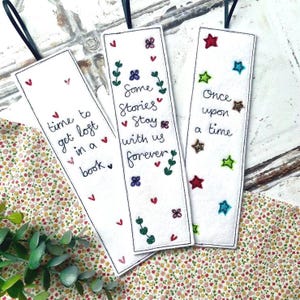 May include: Three white fabric bookmarks with black ribbon ties. Each bookmark has embroidered text and decorative elements. One says "time to get lost in a book," another "Some Stories Stay with us forever," and the last "Once upon a time."
