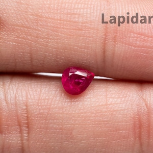 Puede incluir: Una piedra preciosa de rubí facetada en forma de pera con un color rojo intenso. La piedra está colocada entre dos dedos. El texto Lapidarians es visible en la esquina superior derecha de la imagen. El rubí muestra una superficie clara y reflectante.