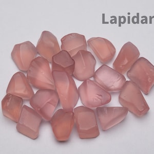 Puede incluir: Una colección de pequeñas piedras pulidas, translúcidas y de color rosa pálido, con formas irregulares y superficies lisas. La palabra Lapidarians está impresa en gris en la esquina superior derecha, sobre un fondo neutro y liso.
