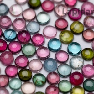 May include: A collection of round, faceted gemstones in various shades of pink, green, blue, and clear. The gemstones are arranged in a random pattern on a white surface.