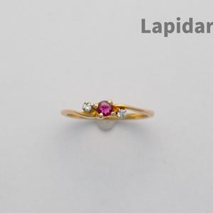 Könnte beinhalten: Ein goldener Ring mit einem zentralen roten Edelstein, flankiert von zwei kleineren, klaren Edelsteinen. Der Ring hat ein zartes, wirbelndes Design. Das Wort "Lapidarians" steht in grauer Schrift über dem Ring.