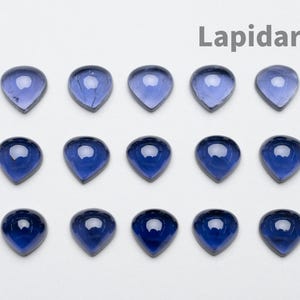 May include: A collection of teardrop-shaped, polished gemstones in shades of blue. The gems are arranged on a white surface, with the word "Lapidarians" above. The stones have a glossy finish.