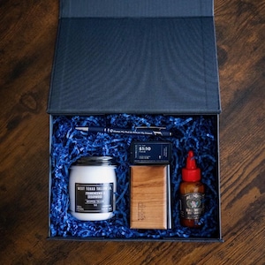 May include: A dark blue gift box filled with blue paper shred containing a West Texas Tallow Co. jar, a blue pen with text, a small black tin labeled 51:10, a wooden Drift car air freshener, and a bottle of Melinda's Black Truffle Hot Sauce.