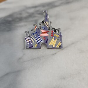 Può includere: Una spilla in smalto color argento a forma di castello. Il castello ha bandiere e striscioni colorati, con uno sfondo grigio e viola. La spilla è su una superficie di marmo.