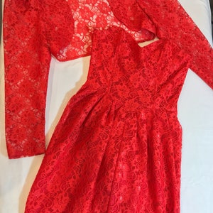 May include: A red lace dress and matching bolero jacket. The dress has a fitted bodice and a full skirt. The bolero has long sleeves and a scoop neckline.
