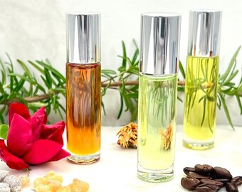 May include: Four glass roller bottles with silver caps filled with different colored liquids. The bottles are arranged in a row on a white surface with various natural elements, including sprigs of rosemary, coffee beans, and dried flowers.