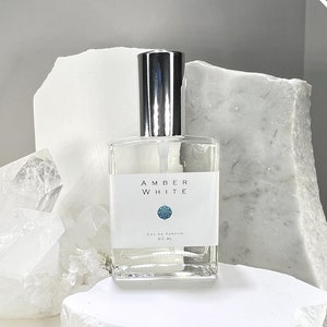 Puede incluir: Una botella de vidrio transparente de Amber White Eau de Parfum, con un pulverizador plateado y una etiqueta blanca con el nombre de la marca y un logotipo azul. La botella está sobre una superficie blanca.