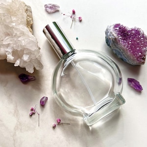 Peut inclure: Un flacon en verre transparent avec un atomiseur argenté et une base ronde et plate. Le flacon est entouré de petits cristaux violets, de fleurs roses et d'un grand amas de cristaux blancs.
