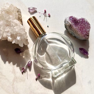 May include: A clear glass bottle with a gold lid and a spray nozzle. The bottle is surrounded by small purple and white crystals and flowers.