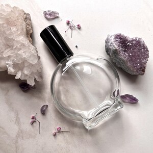 May include: A clear glass bottle with a black spray nozzle, surrounded by amethyst crystals and small pink flowers on a white background.