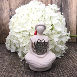 May include: A white ceramic figurine of a seated woman holding a silver flower of life mandala in her hands. The figurine is surrounded by white hydrangeas.