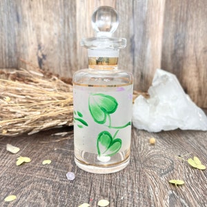 May include: A clear glass bottle with a gold rim and a green leaf design. The bottle has a glass stopper and is filled with a clear liquid.