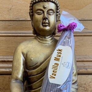 May include: A package of incense sticks with a label that reads "Vanilla Musk 11 inch natural incense". The package is held by a golden statue of Buddha.