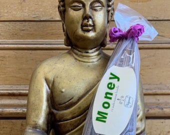 May include: A package of incense sticks with a label that reads "Money" with a green background and a small image of a swirl. The incense sticks are brown and are in a clear plastic bag with a purple ribbon.
